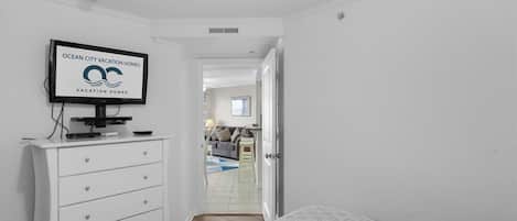 A bedroom with a bed, a TV on a dresser, a white dresser, and a door leading to a living area.
