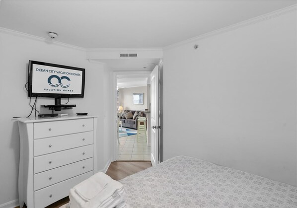 Un dormitorio con cama, televisor sobre una cómoda blanca, y una puerta que da a una sala de estar.