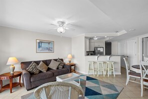 Una sala moderna con un sofá, dos mesas auxiliares, una mesa de centro y un ventilador de techo. Cuenta con una zona de cocina con una barra y taburetes.