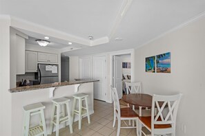 Cocina compacta con gabinetes blancos, encimera de granito y refrigerador de acero inoxidable. Hay dos taburetes blancos para bar y una mesa de comedor de madera con sillas blancas.
