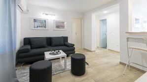 Una sala de estar moderna con un sofá negro, una mesa de centro blanca y dos otomanas negras. Hay dos cuadros enmarcados en la pared, un equipo de aire acondicionado blanco y una silla plegable blanca.