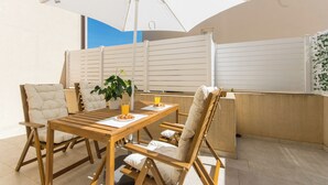 Una mesa de madera con sillas, una planta en maceta y un paraguas blanco en un espacio al aire libre, luminoso y soleado, con vistas al cielo.