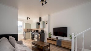 Moderni olohuone, jossa on sohva, sohvapöytä, televisio ja taustalla näkyy keittiö.
