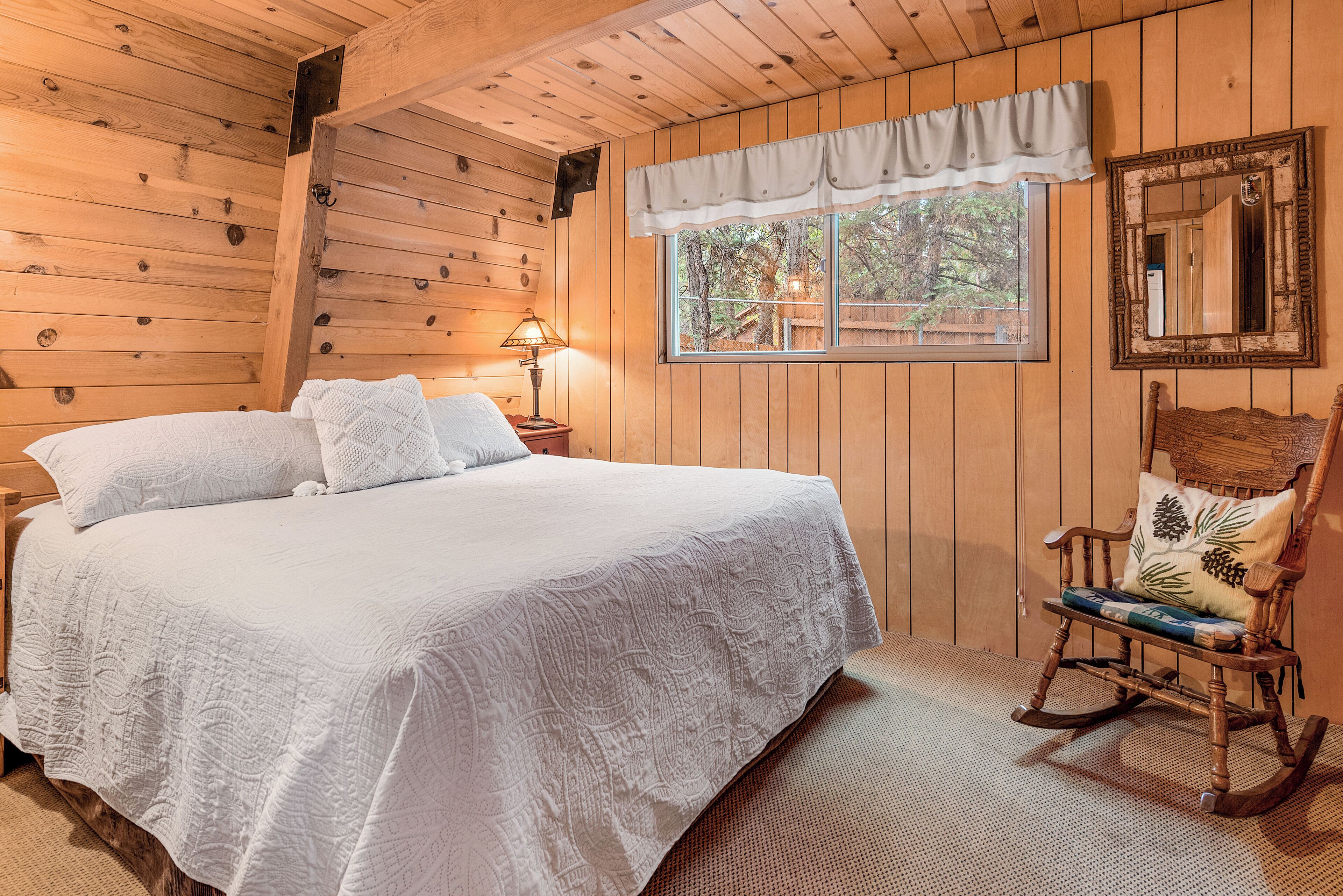 Ein Schlafzimmer einer Holzhütte mit einem großen Bett, einem Schaukelstuhl, einem Fenster mit Blick auf Bäume und einem Spiegel an der Wand.
