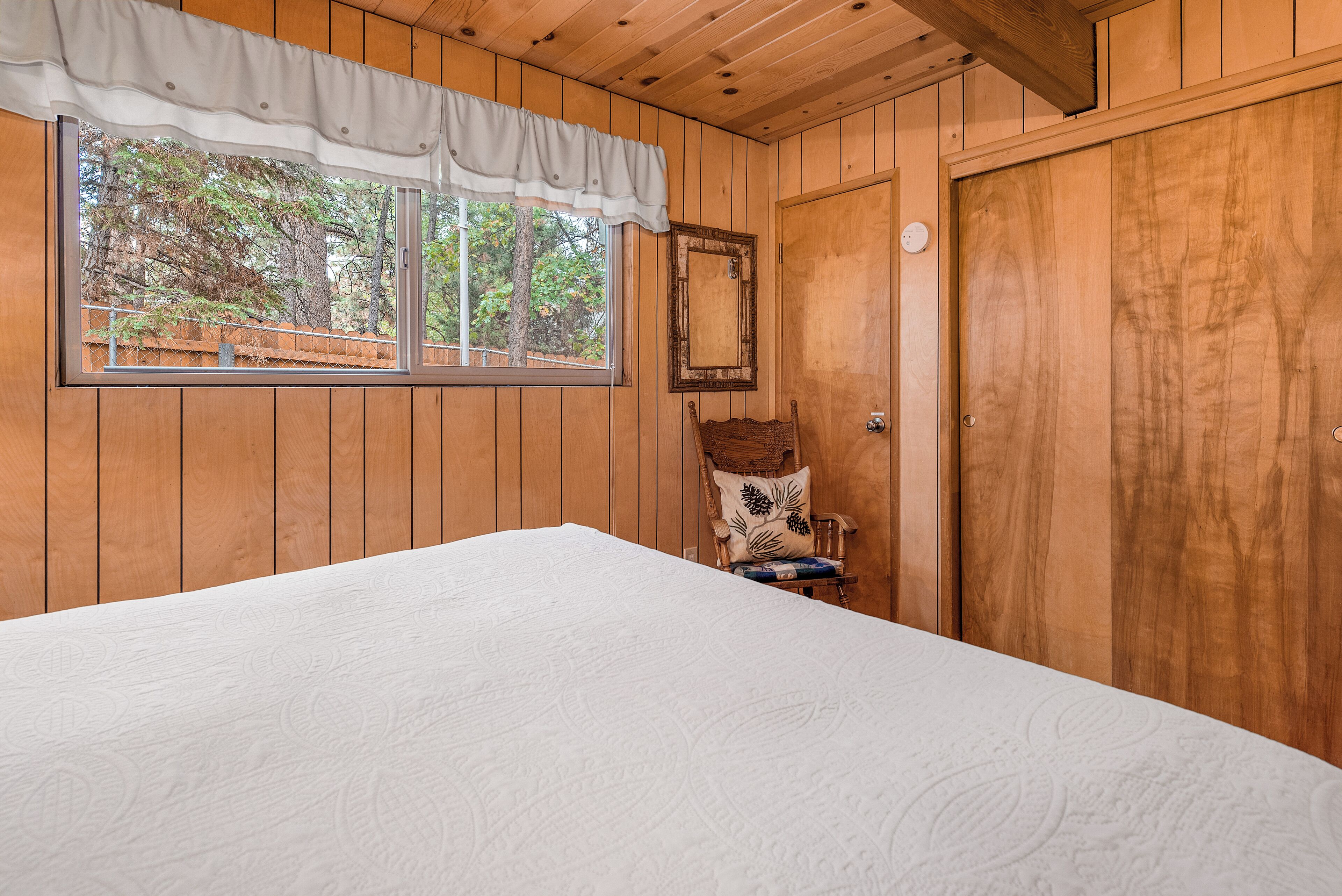 Una habitación con paneles de madera, una cama grande, una silla con cojín, una ventana con cortina y una puerta.