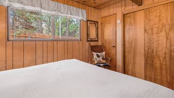Una habitación con paneles de madera, una cama grande, una silla con cojín, una ventana con cortina y una puerta.
