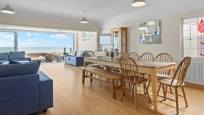 En rummelig opholdsstue med et stort vindue der ud til havet, et spisebord med stole og en blå sofa.