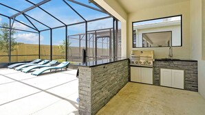 Une véranda avec une structure en verre, un barbecue et un espace au bord de la piscine équipé de chaises longues.