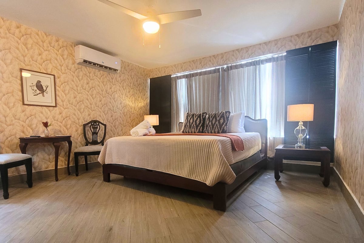 A hotel room with a bed, two bedside tables, a chair, a small table, and a wall-mounted air conditioner.