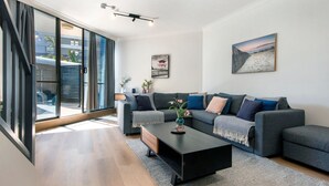 Ein modernes Wohnzimmer mit einem grauen Sofa, einem schwarzen Couchtisch und einem großen Fenster mit Jalousien.