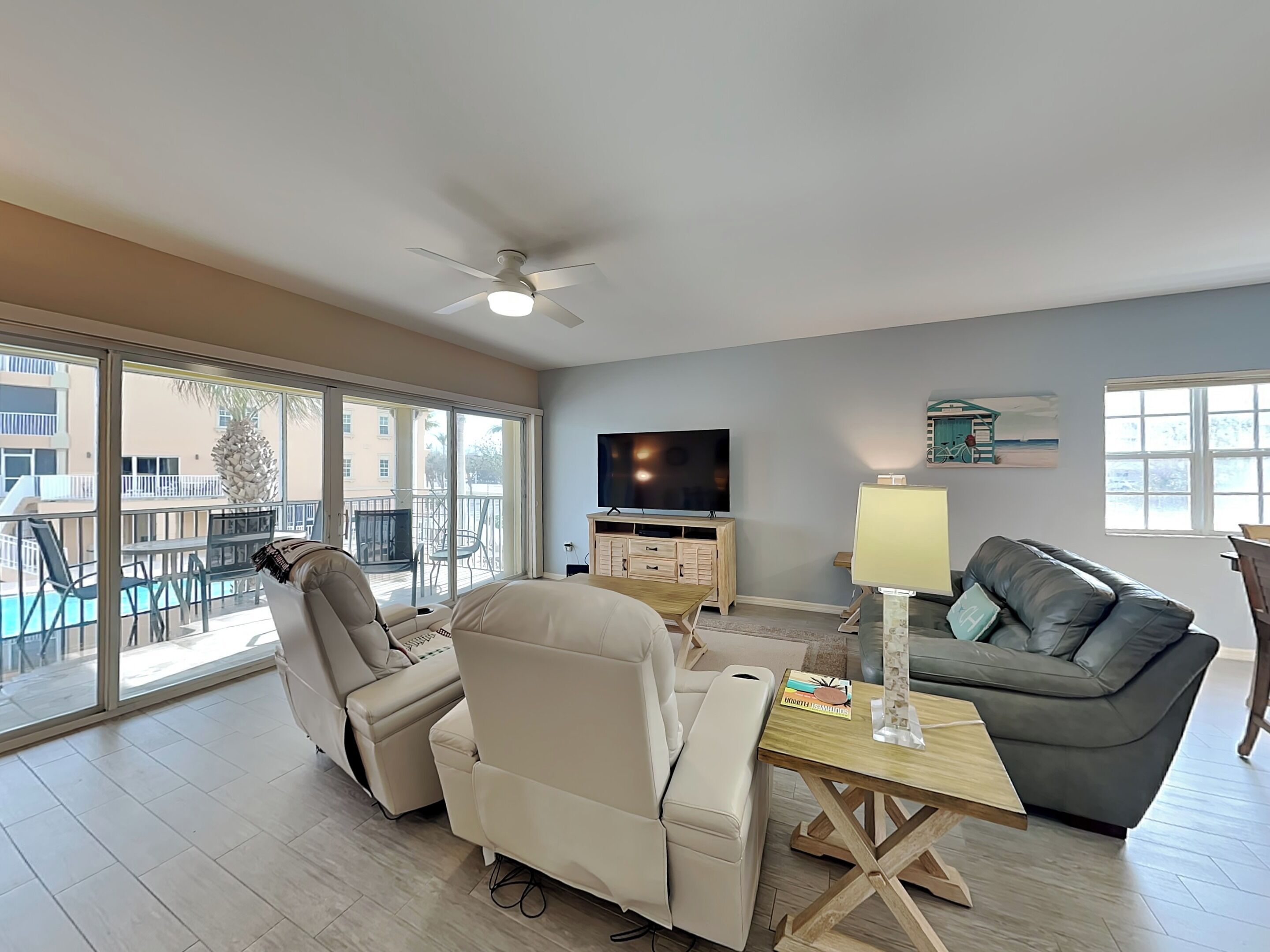 A spacious living room with a large window, a ceiling fan, a flat-screen TV, a grey sofa, and a wooden coffee table.
