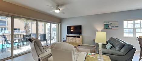 A spacious living room with a large window, a ceiling fan, a flat-screen TV, a grey sofa, and a wooden coffee table.