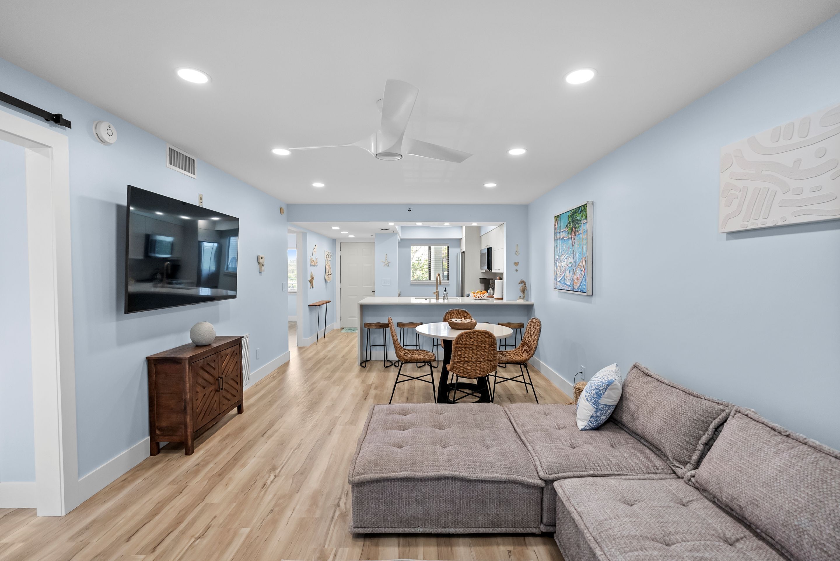 A modern living room with a sectional sofa, a dining area with a round table and chairs, a kitchen with a central island, and a large TV mounted on the wall.