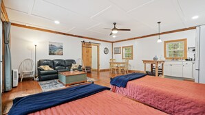 A room with two beds, a sofa, a dining table, a kitchen area, and a ceiling fan.