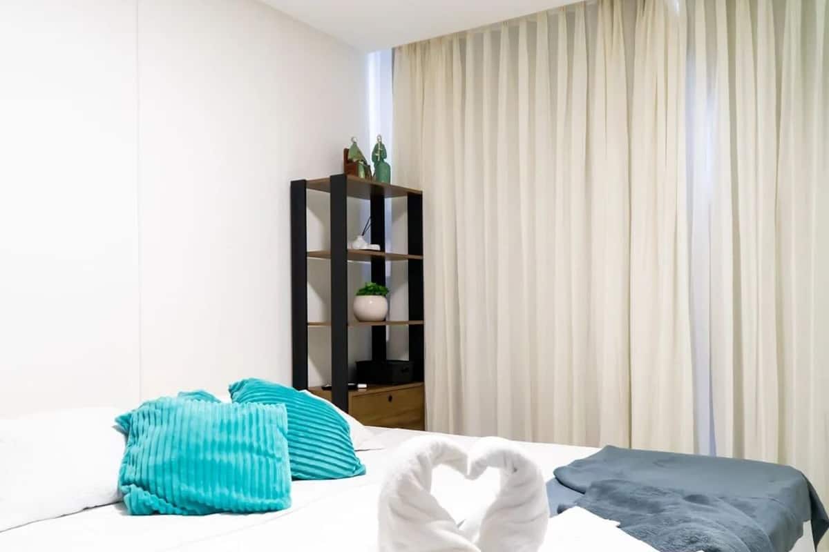 Hotellihuone, jossa on sänky, yöpöytä, kirjahylly ja ikkuna, jossa on verhot.