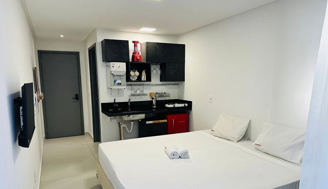 A hotel room with a bed, a desk with a red appliance, a TV, and a door.