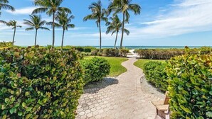A paved pathway leading to a beach with palm trees and lush greenery. - Riviera Club 204 by Distinctive Beach Rentals (Fort Myers Beach)