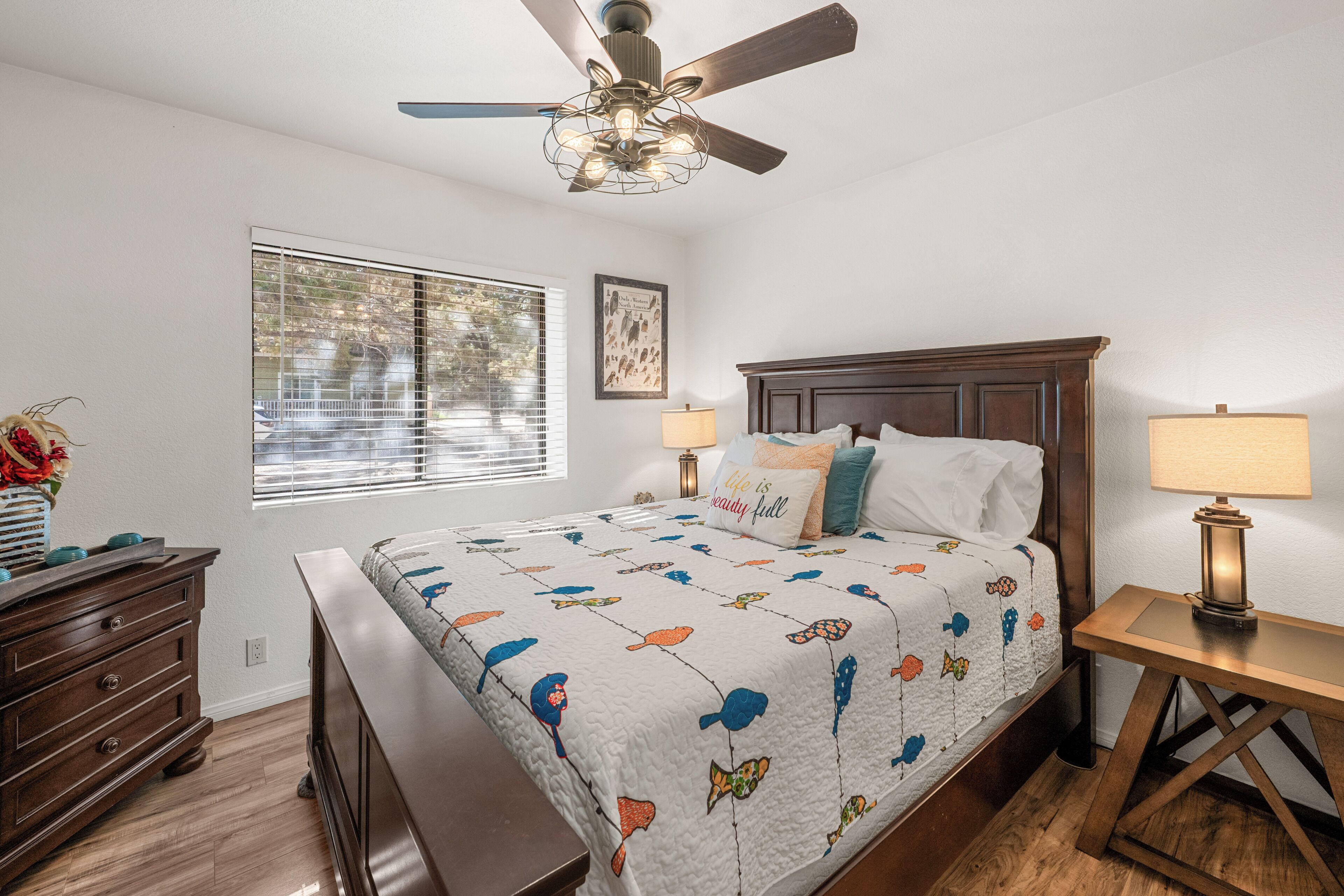 A bedroom with a bed, two bedside tables, a ceiling fan, a window with blinds, and a dresser.