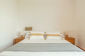 Een netjes opgemaakt bed met witte lakens en twee kussens, een grijze plaid en twee bedlampjes op houten nachtkastjes.