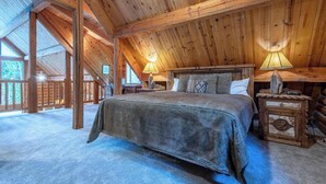Ein gemütliches Schlafzimmer in einer Blockhütte mit einem großen Bett, zwei Nachttischen mit Lampen und Blick ins Freie.