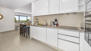 A modern kitchen with white cabinets, a stainless steel sink, and a built-in oven. There are stools at the counter and a window providing a view of the outdoors. - Vista Mar in Quarteira (Loulé)