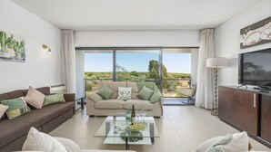 A modern living room with a sofa, coffee table, and a TV. There is a balcony with a view of greenery. - Vista Mar in Quarteira (Loulé)