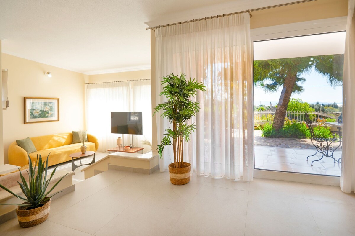 A bright living room with a yellow sofa, a flat-screen TV, and a large window with a view of greenery and a tree.