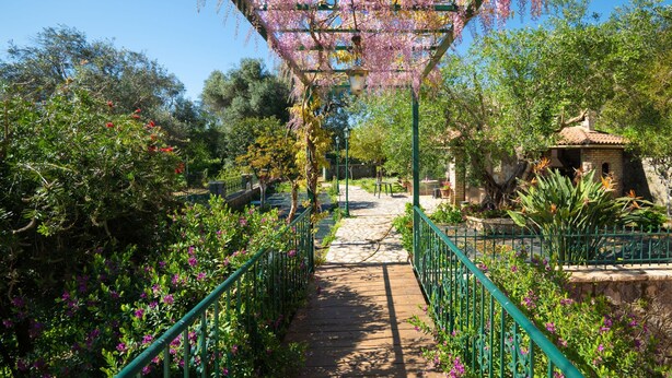 A garden pathway with a pergola covered in pink flowers, leading to a small building with a tiled roof. - Villa Marifei in Kato Korakiana (Corfu)