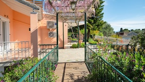 A covered walkway with a wooden floor leading to a building entrance, surrounded by greenery and a pergola with flowering vines. - Villa Marifei in Kato Korakiana (Corfu)