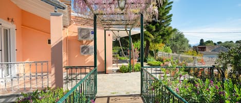 A covered walkway with a wooden floor leading to a building entrance, surrounded by greenery and a pergola with flowering vines.