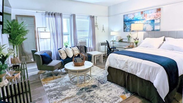 Une chambre d’hôtel moderne avec un grand lit, un bureau, une chaise et un canapé. Il y a une fenêtre avec des rideaux, une plante et un tableau au mur.
