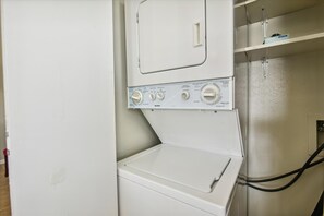 A front view of a white washing machine with control panel, a dryer, and a shelf above the dryer. - Summer Daze by Ryson Vacation Rentals (Galveston)