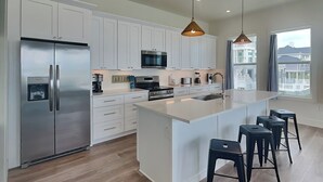 A modern kitchen with white cabinetry, stainless steel appliances, a large island with bar stools, and pendant lighting. - Casa Abide by Ryson Vacation Rentals (Galveston)