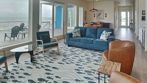 A spacious living area with a large window overlooking the sea, a blue sofa, and a patterned rug. - Casa Abide by Ryson Vacation Rentals (Galveston)