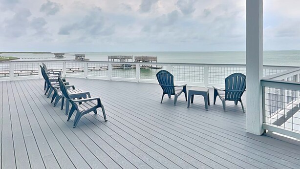 A wooden deck with chairs and tables, overlooking a body of water and a cloudy sky. - Casa Abide by Ryson Vacation Rentals (Galveston)