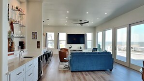 A modern kitchen with white cabinets and a dining area with a blue sofa. There is a large TV and a ceiling fan in the living area. - Casa Abide by Ryson Vacation Rentals (Galveston)