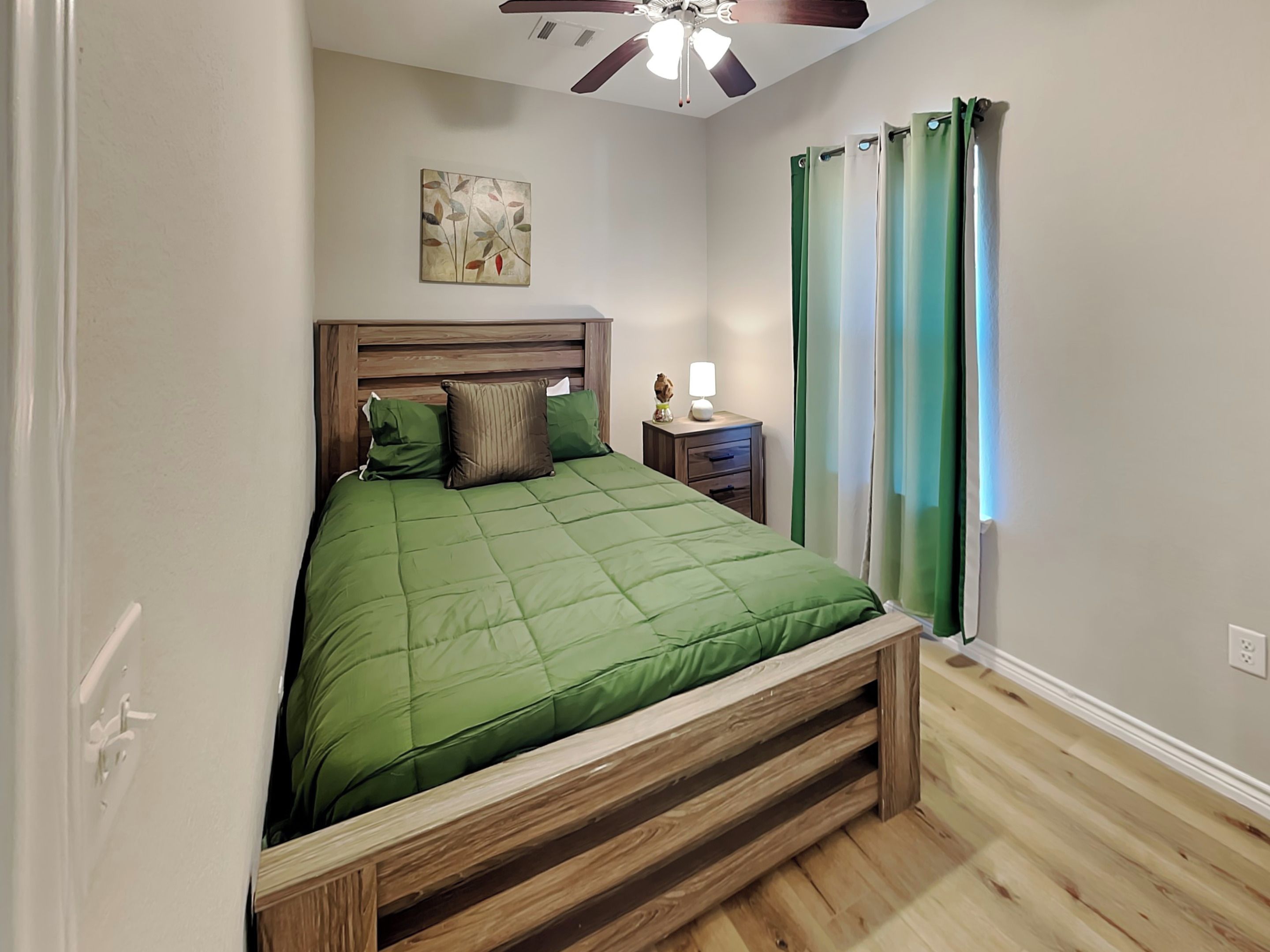 A bedroom with a wooden bed frame, green bedding, a nightstand with a lamp, and a ceiling fan.