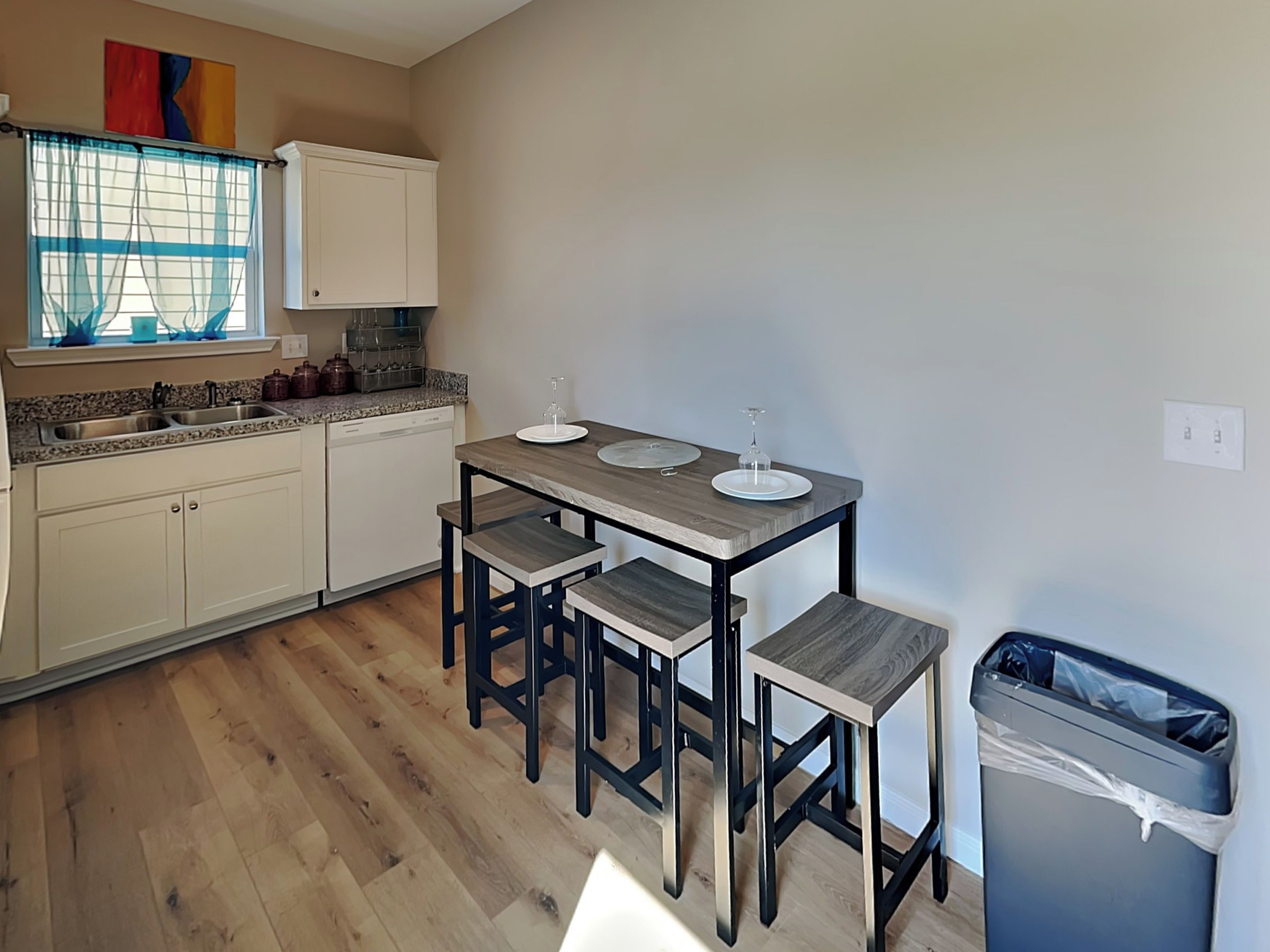 Una cocina con una mesa de madera, cuatro sillas, un lavamanos, armarios y un cubo de basura.