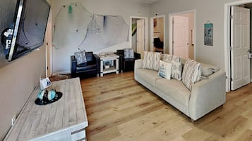 A cozy living room with a wooden coffee table, a sofa with patterned cushions, a TV mounted on the wall, and a ceiling fan.
