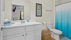 A bathroom with a white sink, mirror, and toilet. There is a framed picture on the wall and a blue shower curtain. - Beach Ballin' by Ryson Vacation Rentals (Galveston)