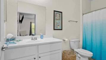 Un baño con lavamanos blanco, espejo y inodoro. Hay un cuadro en la pared y una cortina de ducha azul.
