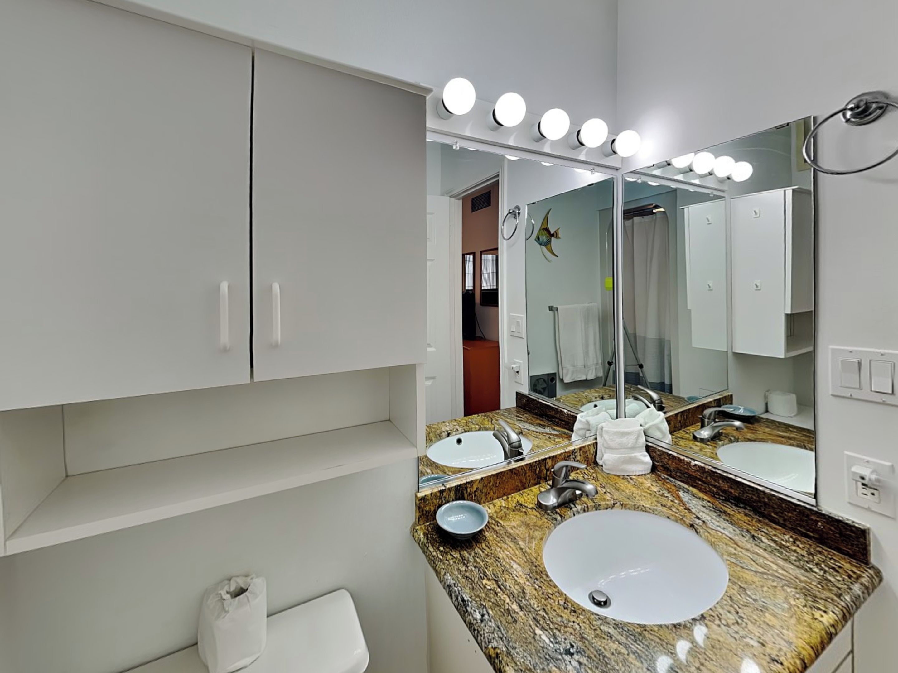 A bathroom with a vanity, two sinks, a mirror with lights, and a toilet.