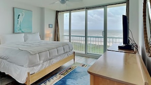 Un dormitorio con cama, ventilador de techo, televisor y una ventana grande con vista a la playa.