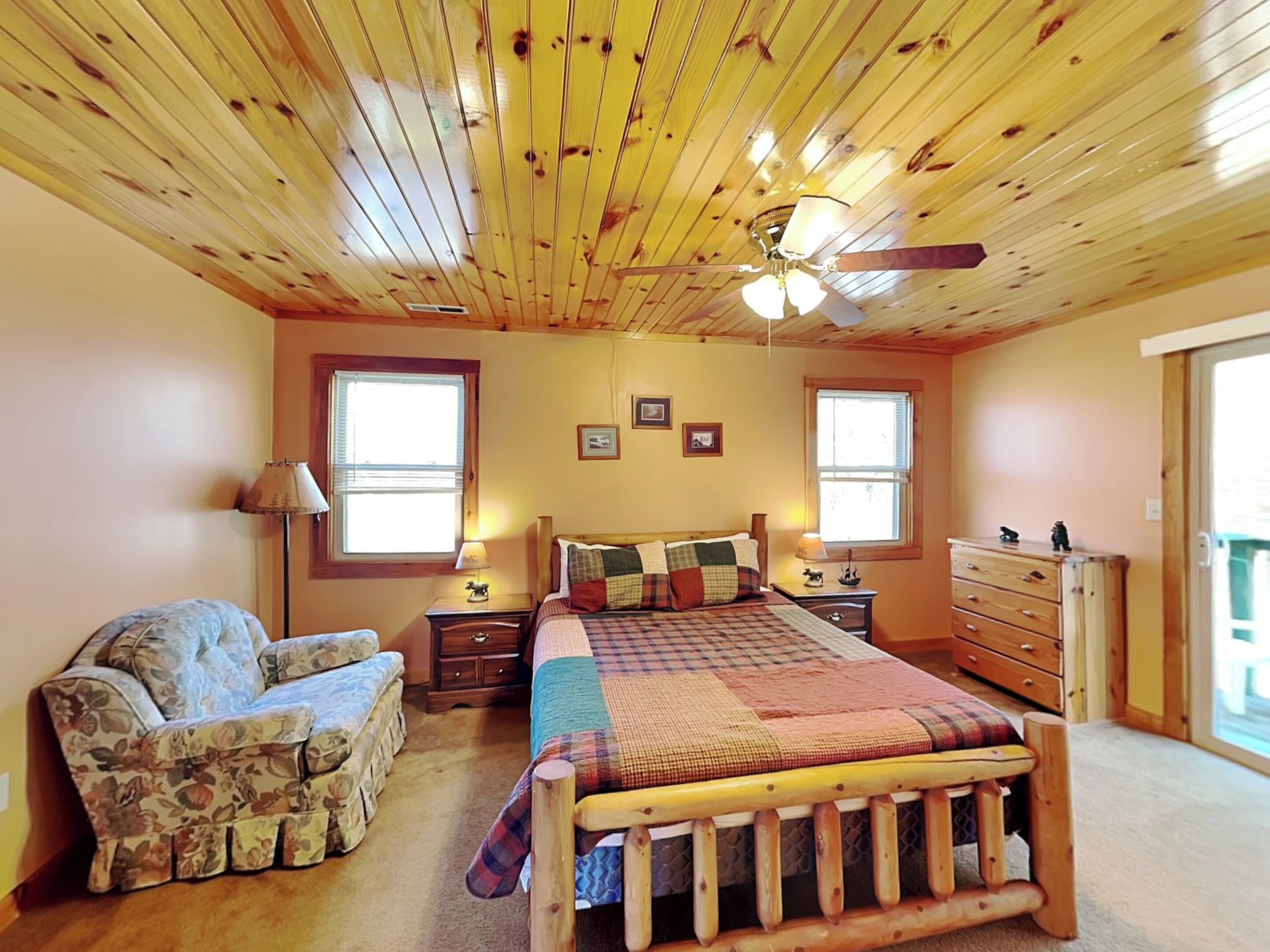 A cozy bedroom with a wooden ceiling, a bed with a plaid bedspread, a floral armchair, and a dresser.