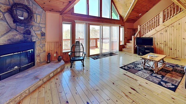 Une pièce spacieuse dotée d’une cheminée en pierre, de planchers en bois, d’un ventilateur de plafond et de grandes fenêtres offrant une vue sur l’extérieur.