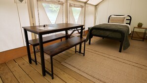 Een tent met een houten tafel en bank, een bed met een groene dekbedovertrek, en een tafeltje met een plant.