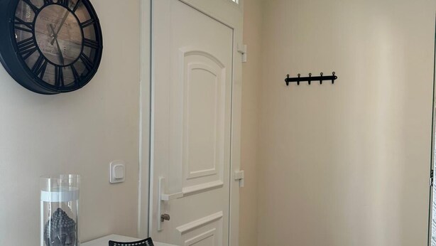 Un pasillo con una puerta blanca, un reloj fijado a la pared, un estante pequeño con un recipiente de vidrio y un objeto negro, y un perchero negro montado en la pared.