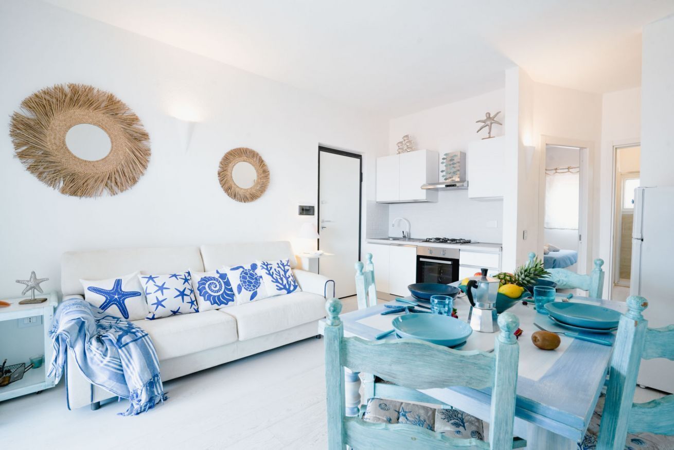 A modern living room with a white sofa, blue and white patterned cushions, a dining table with blue and white chairs, and a kitchen in the background.