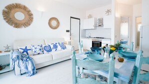 Un salón moderno con un sofá blanco, cojines estampados en azul y blanco, una mesa de comedor con sillas azules y blancas, y una cocina al fondo.