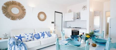 A modern living room with a white sofa, blue and white patterned cushions, a dining table with blue and white chairs, and a kitchen in the background.